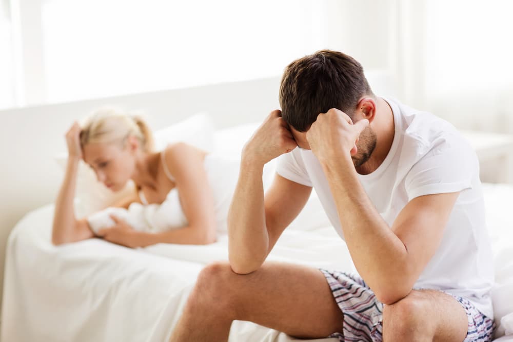 Frustrated guy sitting in a bed while girl lays in bed staring into empty space